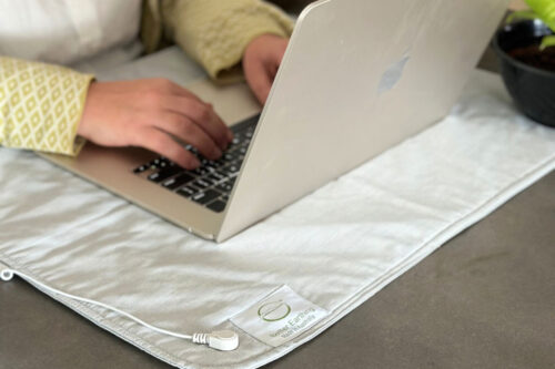 Person typing on a laptop placed on a light gray Better Earthing Fabric Grounding Mat, showcasing its use for grounding while working.