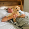 Person resting in bed with a dog on their chest while zipped inside the Better Earthing Recovery Bag, showing how it’s used for grounding and EMF shielding during sleep.